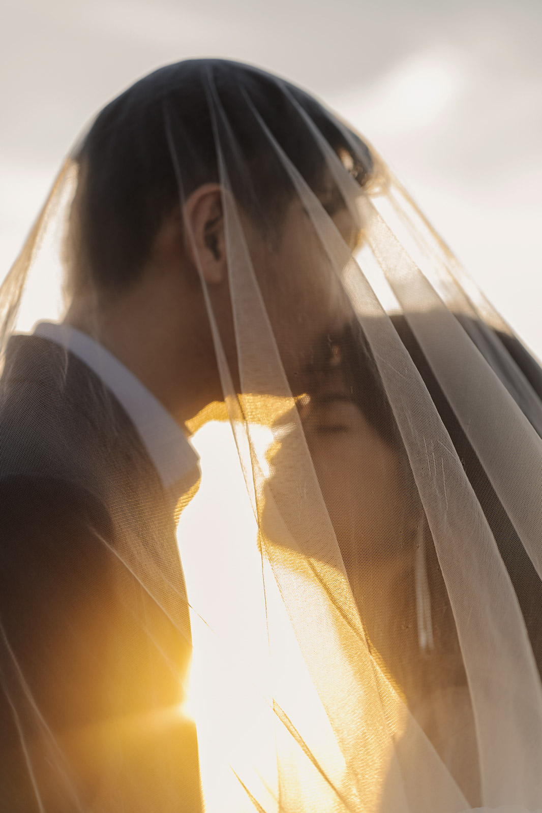 新人在薄紗下接吻,逆光夕陽營造柔暖婚紗情境
