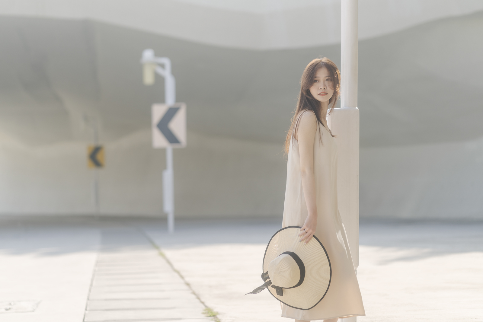 女孩靠著白色燈柱側身站立,手持草帽看向鏡頭