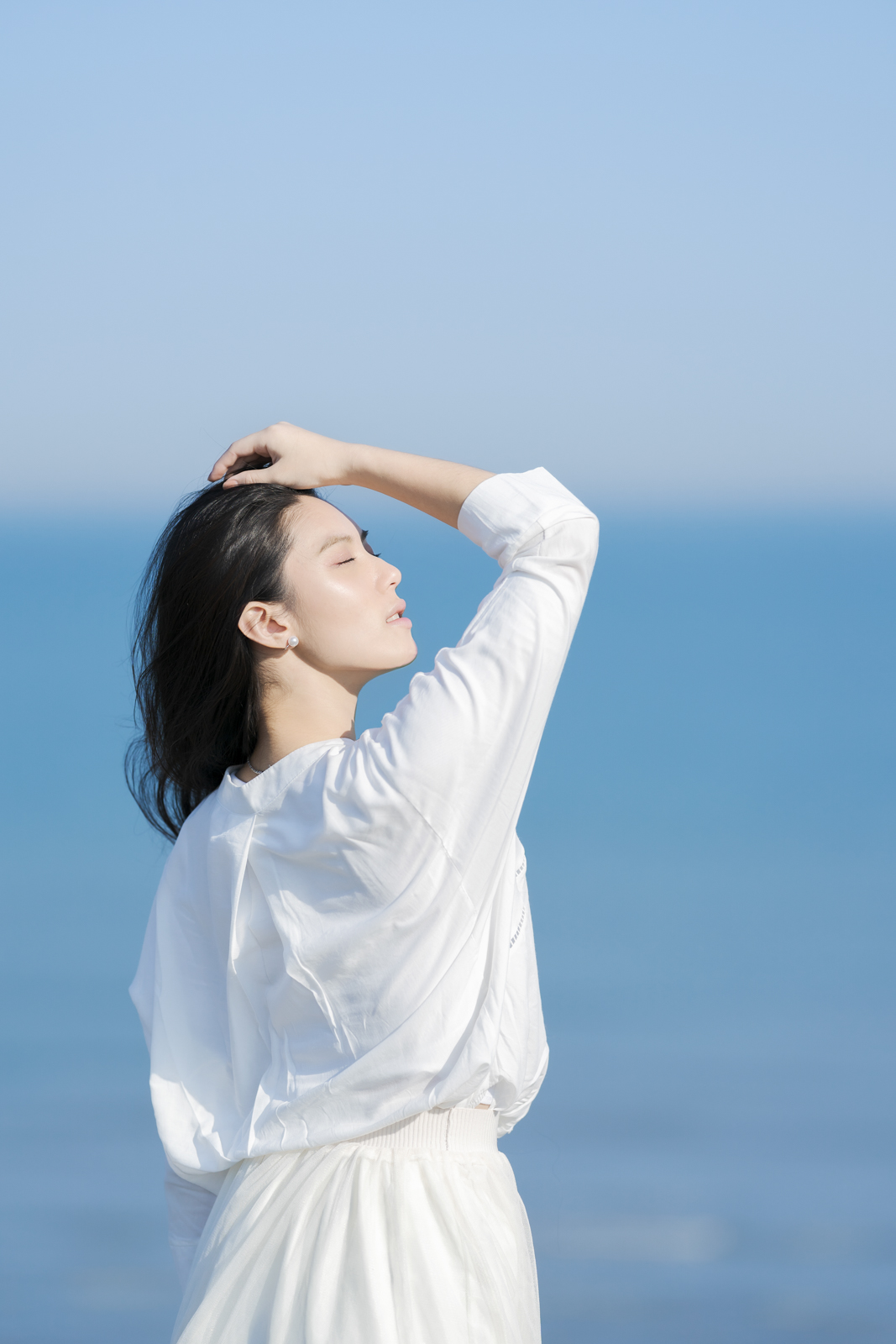 模特面朝陽光閉眼輕抬手臂,背景是柔和漸層的藍色海洋