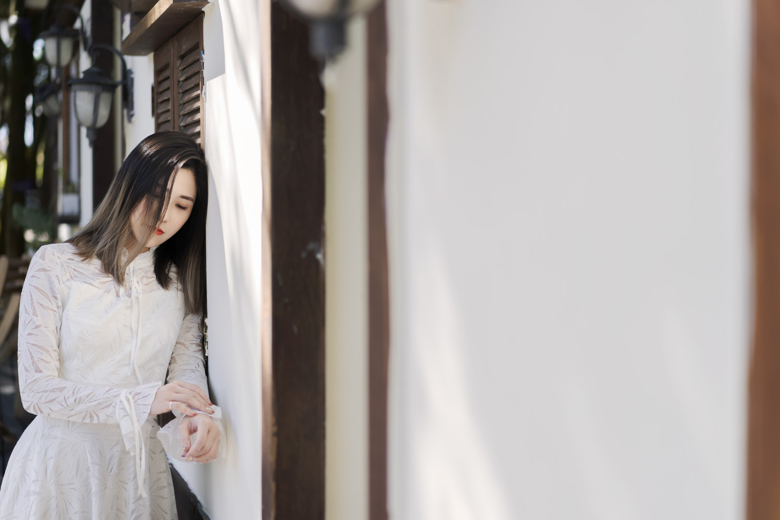身穿白洋裝的女子倚靠木窗牆邊,光線灑落在她的頭髮與袖口