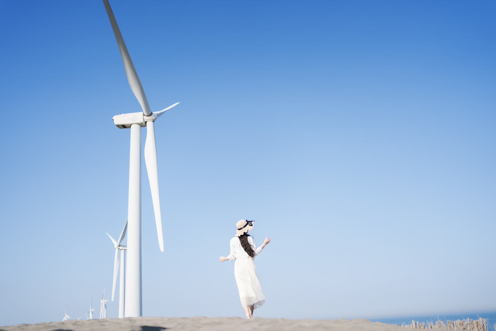 模特站在風力發電機前的沙丘上,身穿白裙望向遠方,背景是晴朗藍天