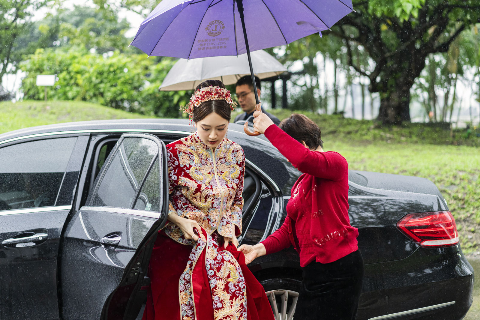 新娘穿著中式禮服在雨中下車，親人撐傘協助
