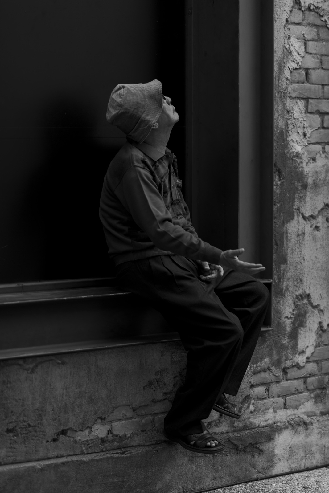 街邊老人在磚牆前抬頭伸手，黑白紀實場景