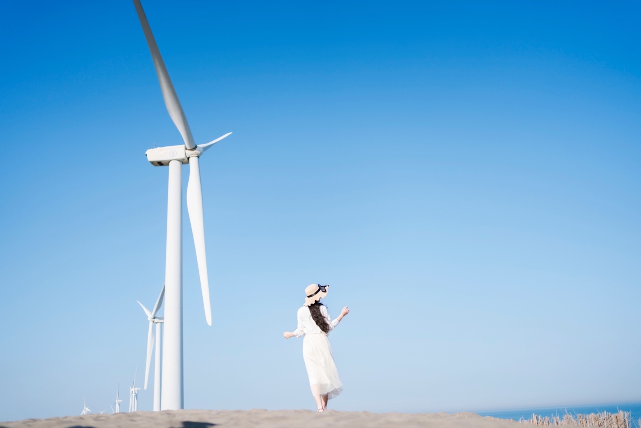 模特站在風力發電機前的沙丘上，身穿白裙望向遠方，背景是晴朗藍天