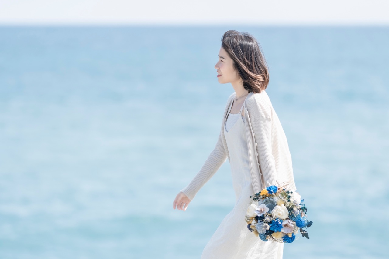 拿著花束的女子微笑側身走在海邊
