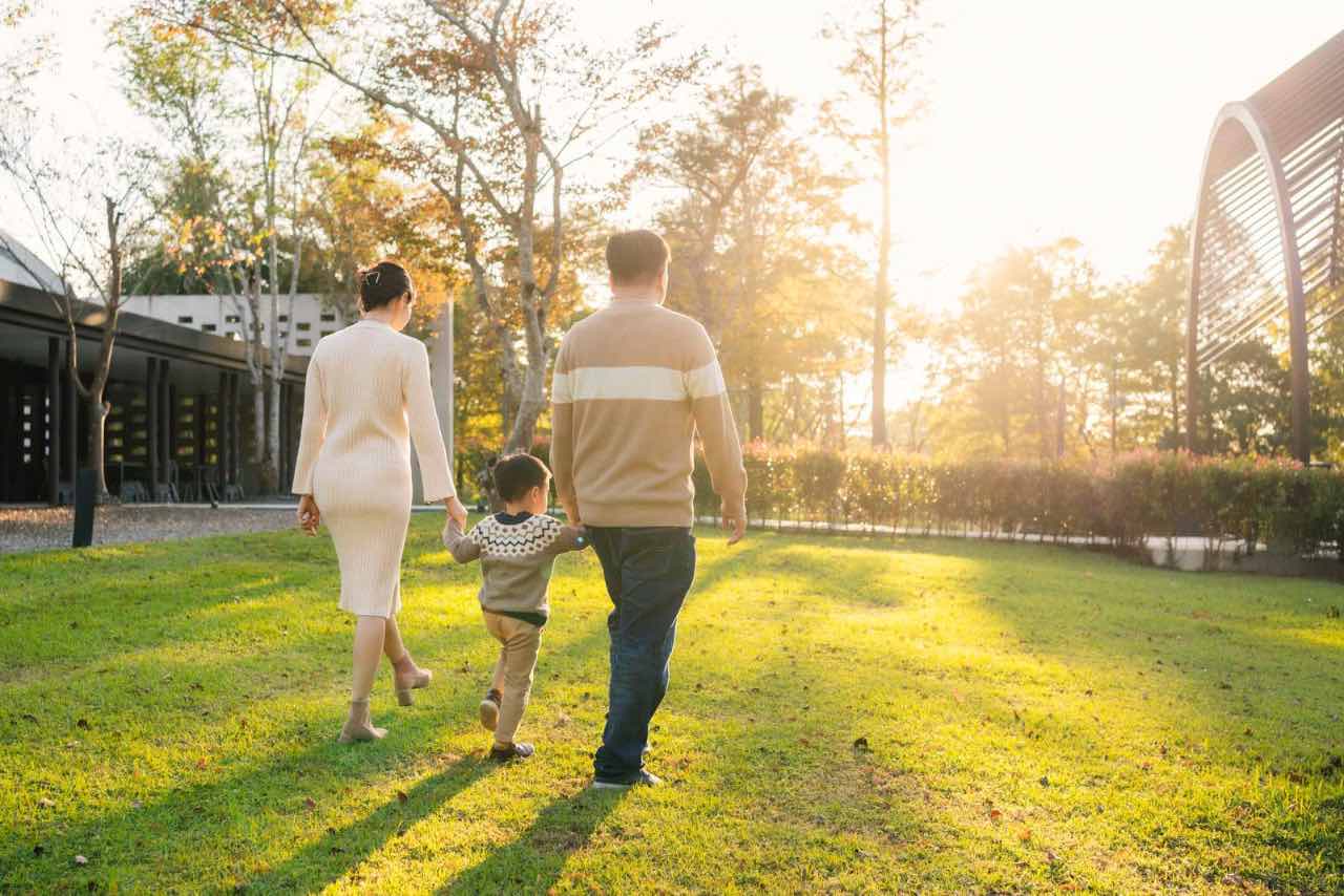 孕婦牽著孩子和先生走在草地上,逆光下步伐一致,畫面溫暖