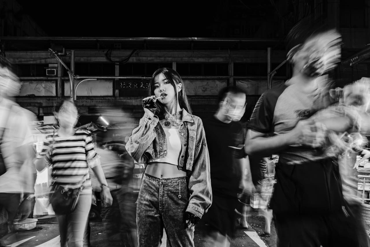 夜市中女孩凝視鏡頭，周圍人群動態模糊黑白對比