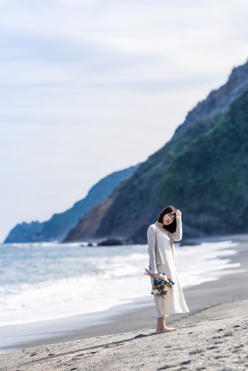 身穿白洋裝的女子站在斜坡沙灘上,手持花束低頭思考