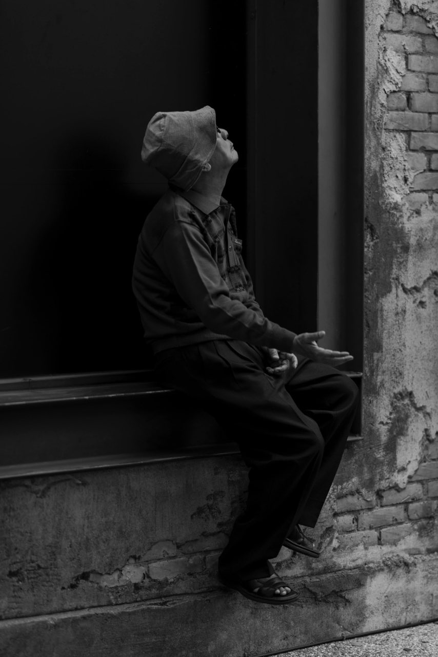 街邊老人在磚牆前抬頭伸手，黑白紀實場景
