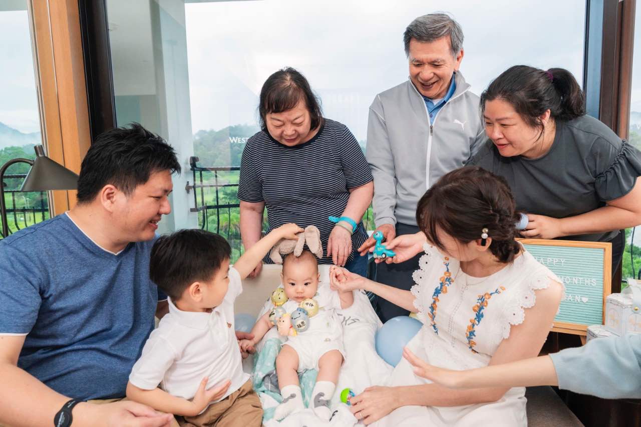 家族成員在房內圍坐互動，嬰兒成為大家目光與動作的中心，氣氛歡樂