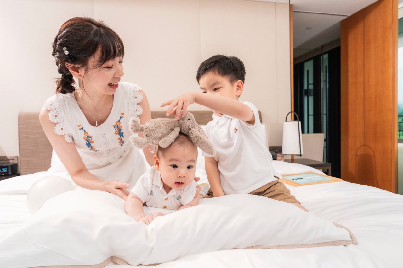媽媽在床上陪伴兩位孩子，哥哥把玩偶放在嬰兒頭上互動嬉戲
