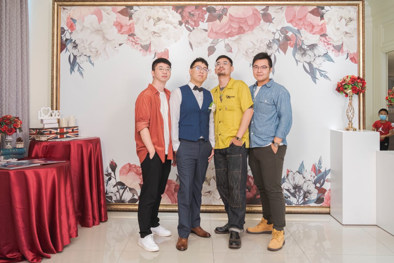 婚禮現場四位好友合照，背景是花田盛事會館的花卉佈景，右上角可見一位穿紅衣的小男孩。