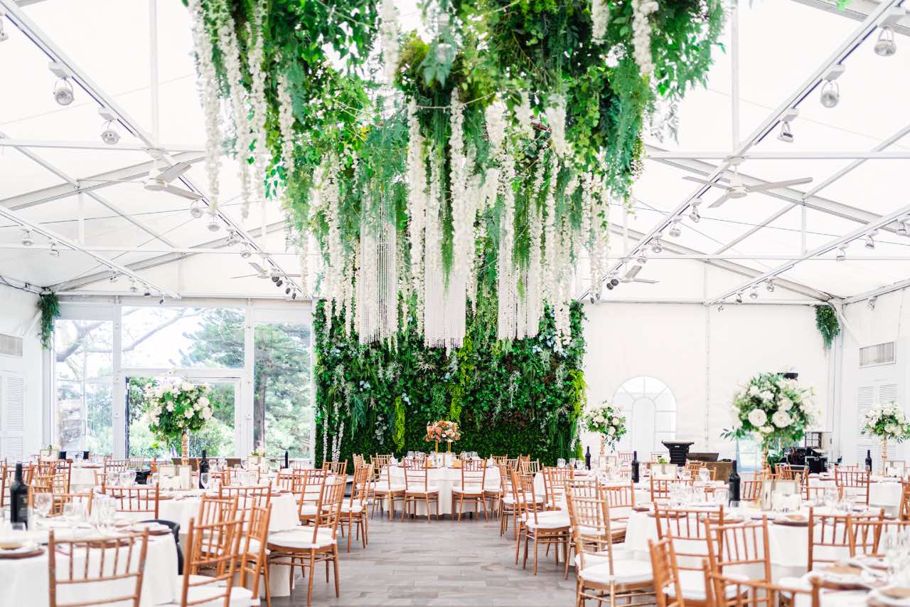 淡水嘉廬婚禮宴會場佈景，白色帳篷天花板與綠植垂掛裝飾，整體光線明亮自然，桌花與木椅呈現典雅氛圍。