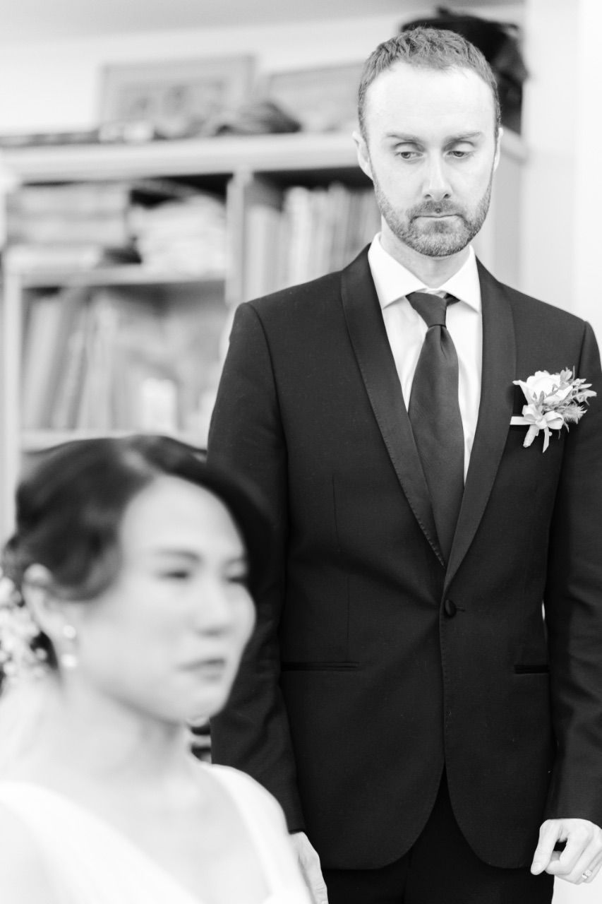 黑白照片中，外國新郎穿西裝配胸花站在室內，深情注視前方正在拜別的新娘，新娘在前景略為失焦。