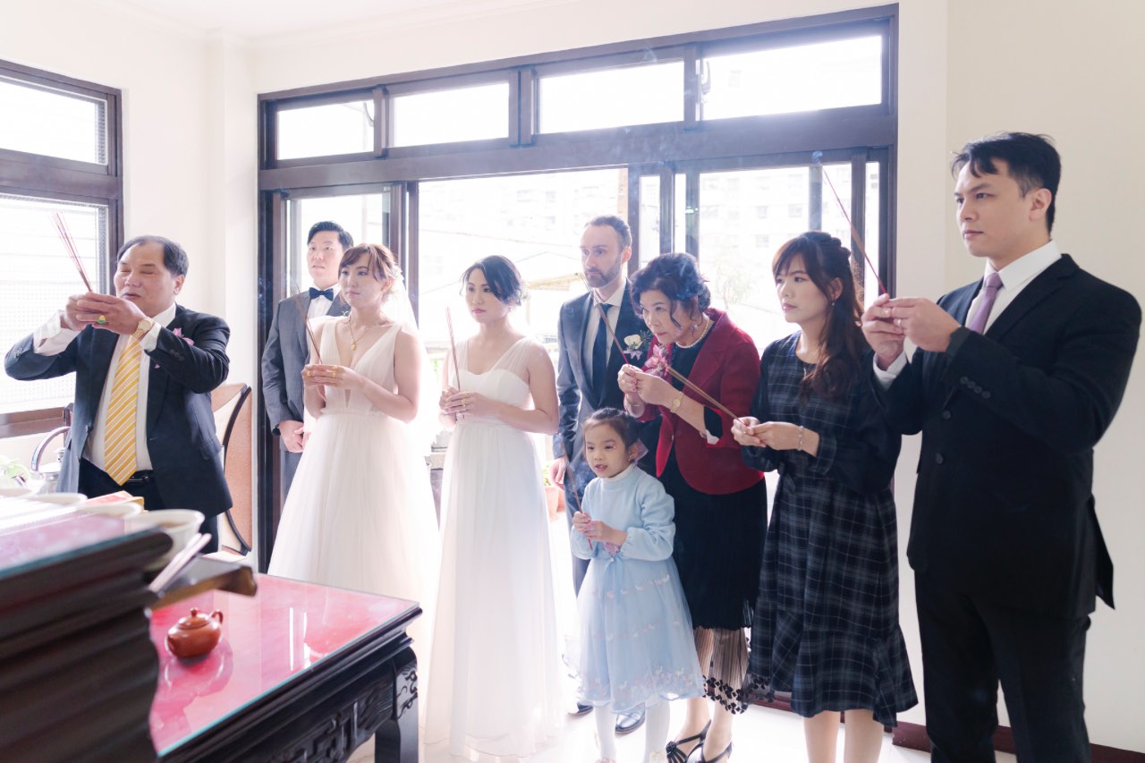 新人與家人站在祖先牌位前手持線香祭祖，長輩帶領儀式，親友一同參與，室內窗邊光線明亮。