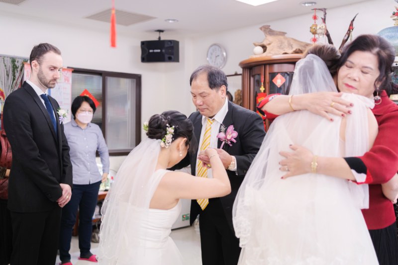 兩位穿白紗的新娘姐妹向父母拜別，一位新娘蹲跪在父親前方行禮，另一位與母親擁抱道別，外國新郎在旁安靜觀看。