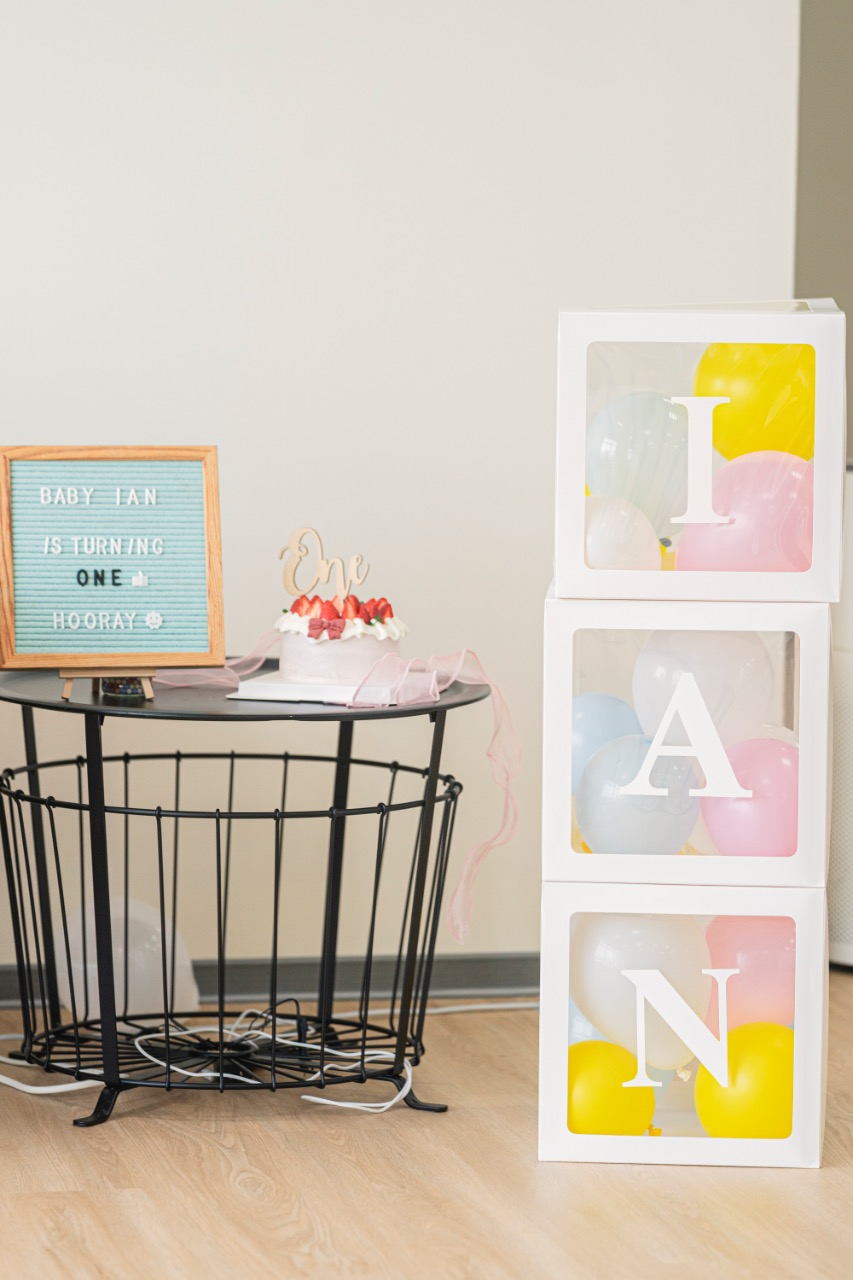 黑色小桌上擺著周歲小蛋糕與字母擺飾，左側有英文字母板寫著寶寶滿一歲祝賀語，右側透明盒疊出寶寶英文名字字母並放入彩色氣球。