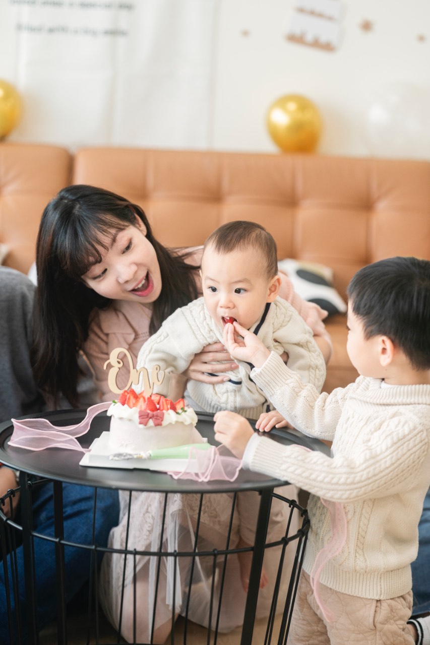 媽媽抱著抓周主角靠近周歲蛋糕，哥哥站在右側伸手餵寶寶吃蛋糕，桌上有草莓蛋糕與「one」字樣裝飾，背景是沙發與氣球佈置。