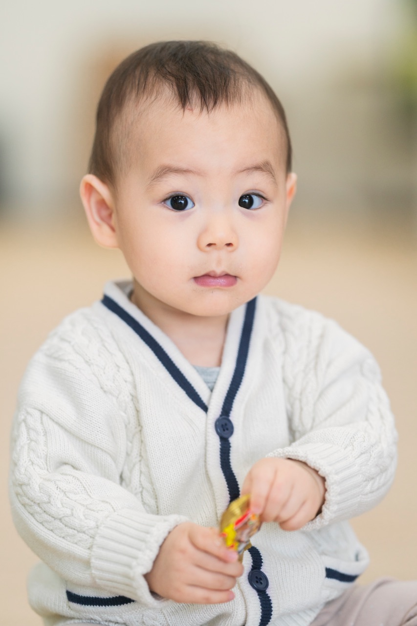 穿白色針織外套的寶寶看向鏡頭，雙手拿著小玩具，背景柔焦呈現室內自然光氛圍。