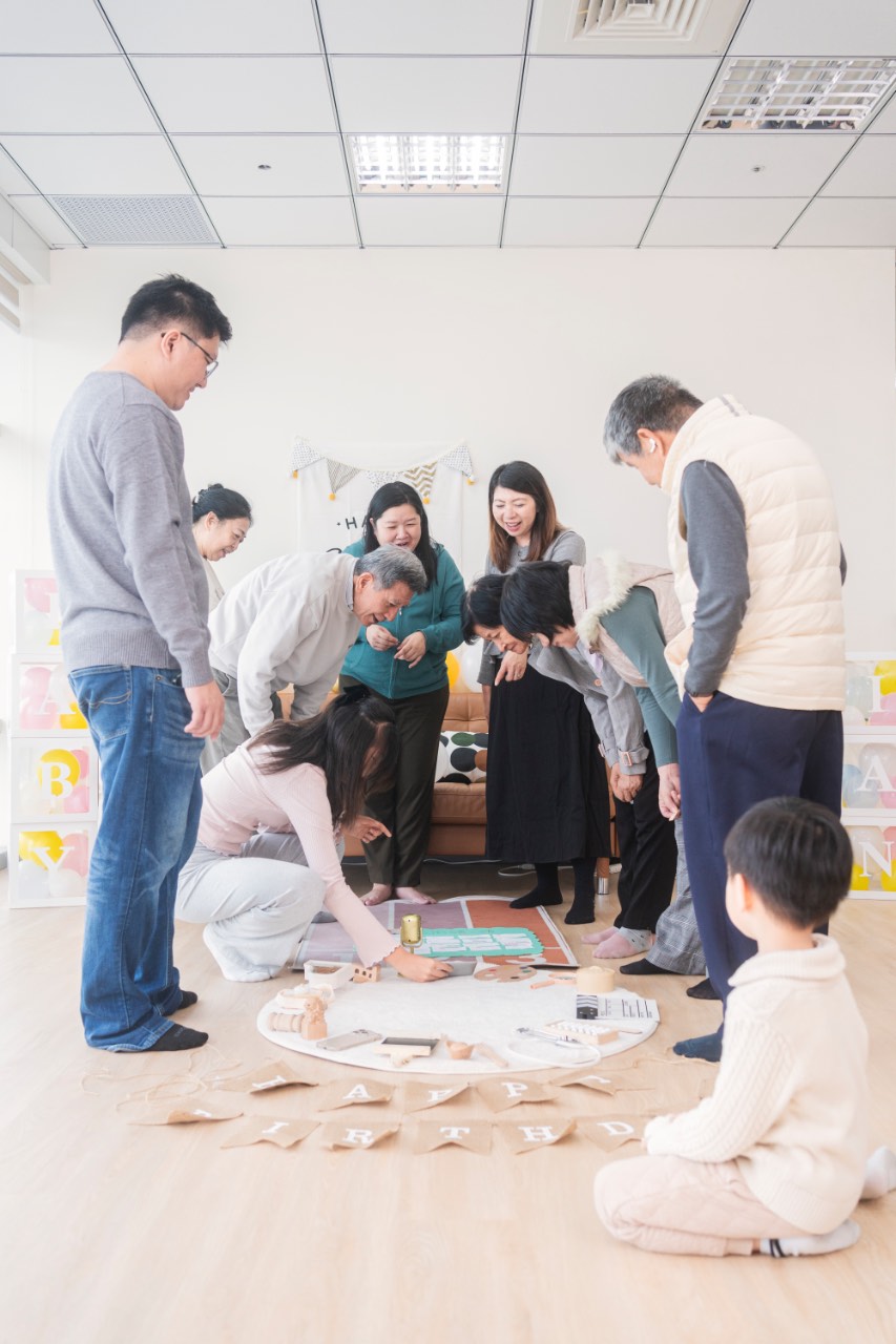 多位親友圍在客廳地上低頭討論並貼紙下注，中央放著抓周猜道具遊戲板與周歲佈置，旁邊有孩子坐著觀看。