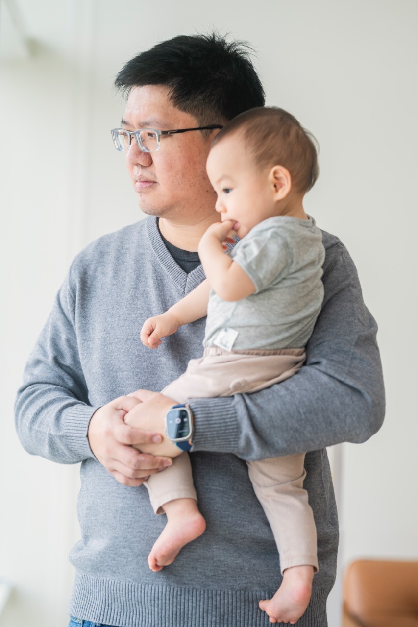爸爸抱著寶寶站在窗邊，兩人望向窗外，寶寶咬著手指，背景明亮柔和呈現自然光氛圍。