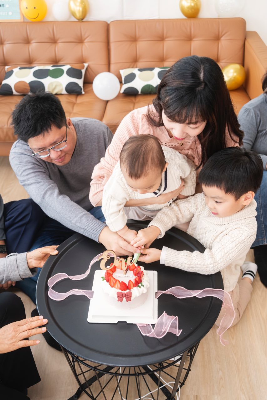 家人圍在黑色圓桌旁一起切周歲蛋糕，媽媽抱著寶寶靠近蛋糕，旁邊孩子握著刀，桌上有草莓蛋糕與「one」字樣裝飾。