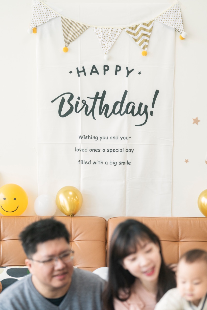 牆面掛著「Happy Birthday」布旗與三角彩旗，沙發上方有金色與白色氣球，前景人物模糊帶出周歲派對氛圍。
