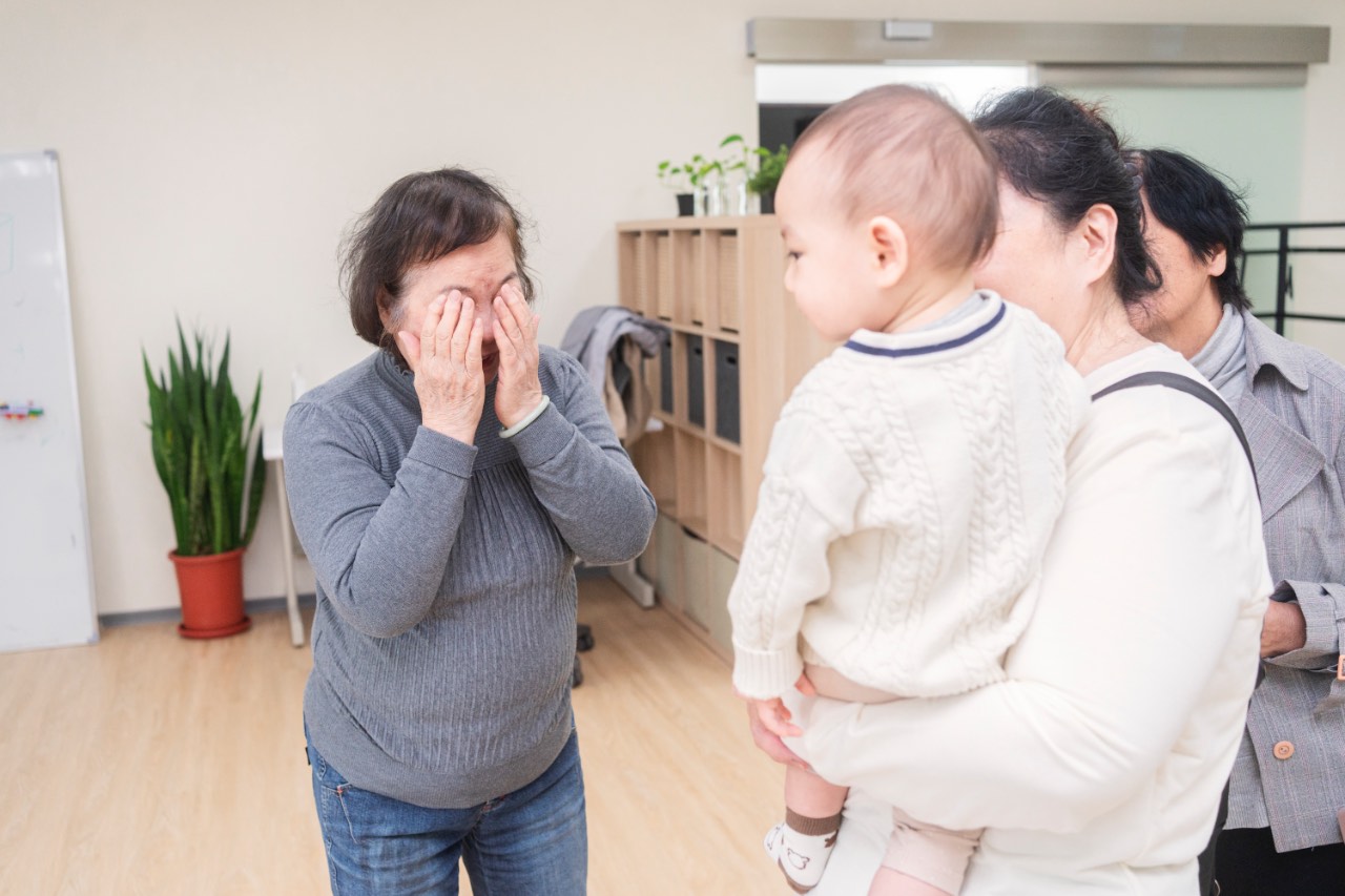 外婆用雙手遮住臉逗小寶寶，奶奶抱著穿白色毛衣的孩子，家人在旁邊圍觀互動的抓周拍攝現場。