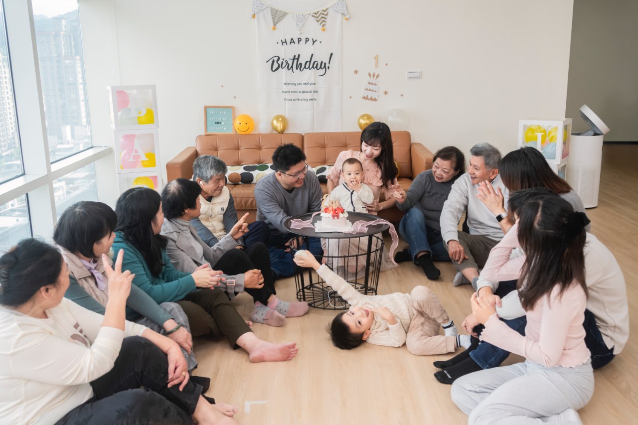 一家人圍坐在客廳地上看著寶寶與蛋糕，背景有「Happy Birthday」佈置，現場多人互動熱鬧，前景有小孩躺在地上。