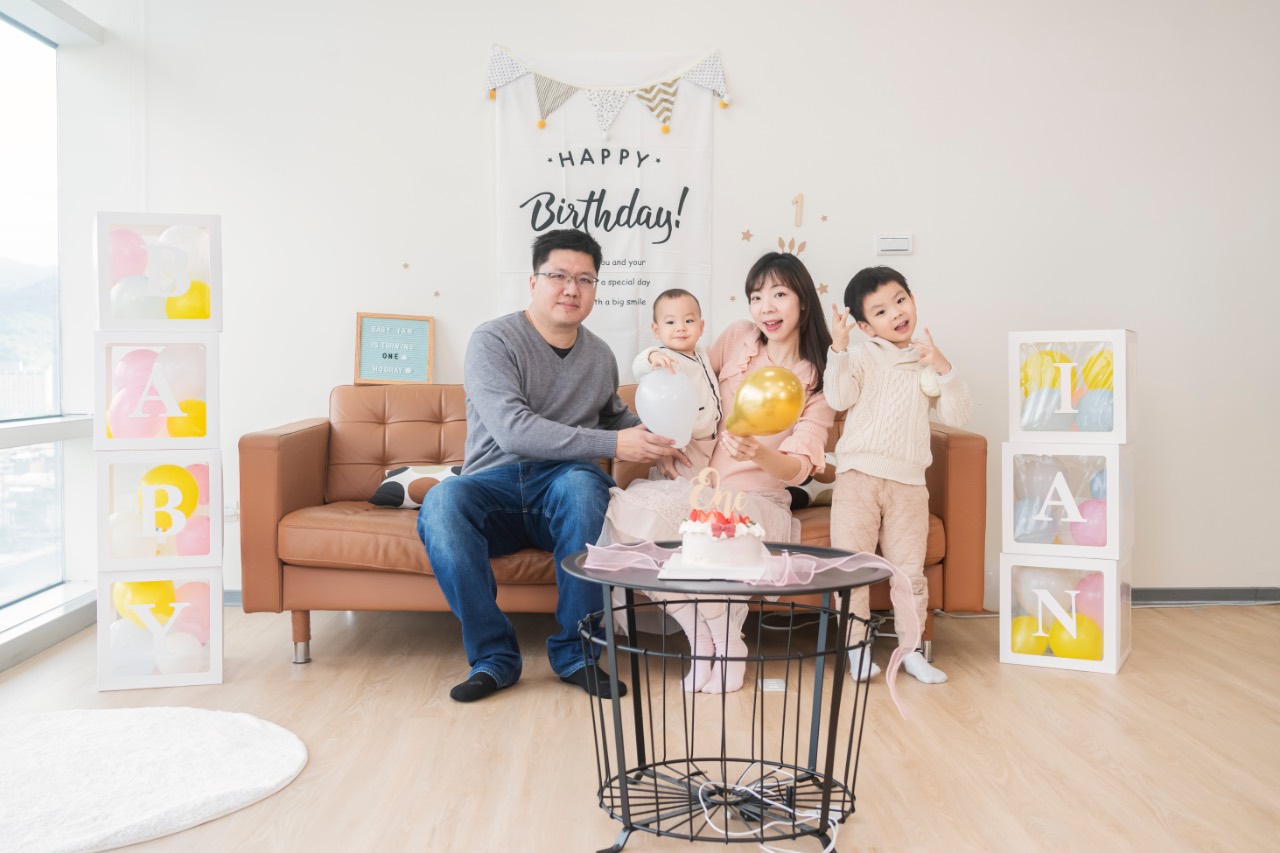 爸爸坐在沙發旁，媽媽抱著寶寶，旁邊有另一位孩子比手勢，前方桌上有蛋糕與氣球，背景是生日佈置與字母積木櫃。