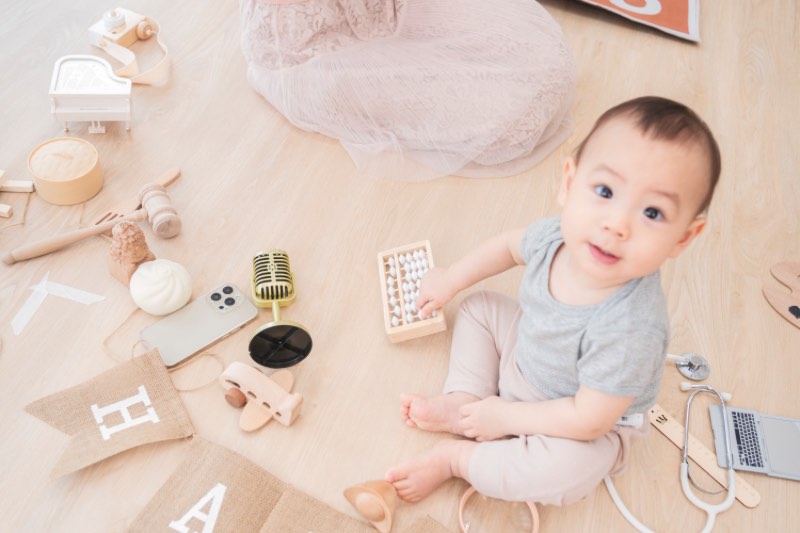 寶寶坐在木地板上抬頭看鏡頭，身旁散落抓周道具與玩具，包含木製小物、麥克風道具與小道具配件。
