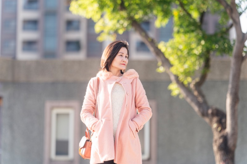 女性站在城市戶外，穿粉色大衣，雙手插口袋，視線望向左側遠方，背景為建築與樹木虛化