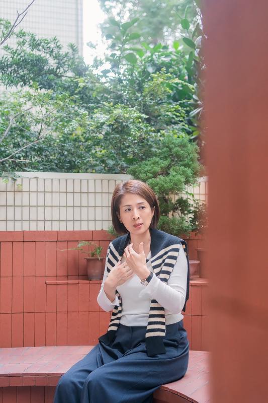 BabyFace 喜餅老闆娘坐在紅磚牆邊的戶外空間，雙手交疊，神情專注地說話，背景為綠色植栽